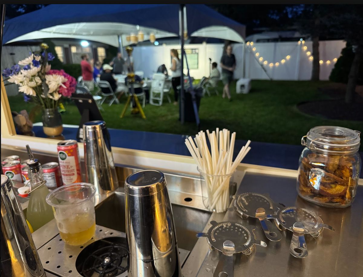 View from behind the Compass bar top with shakers and strainers, looking out at the draft party under the tent
