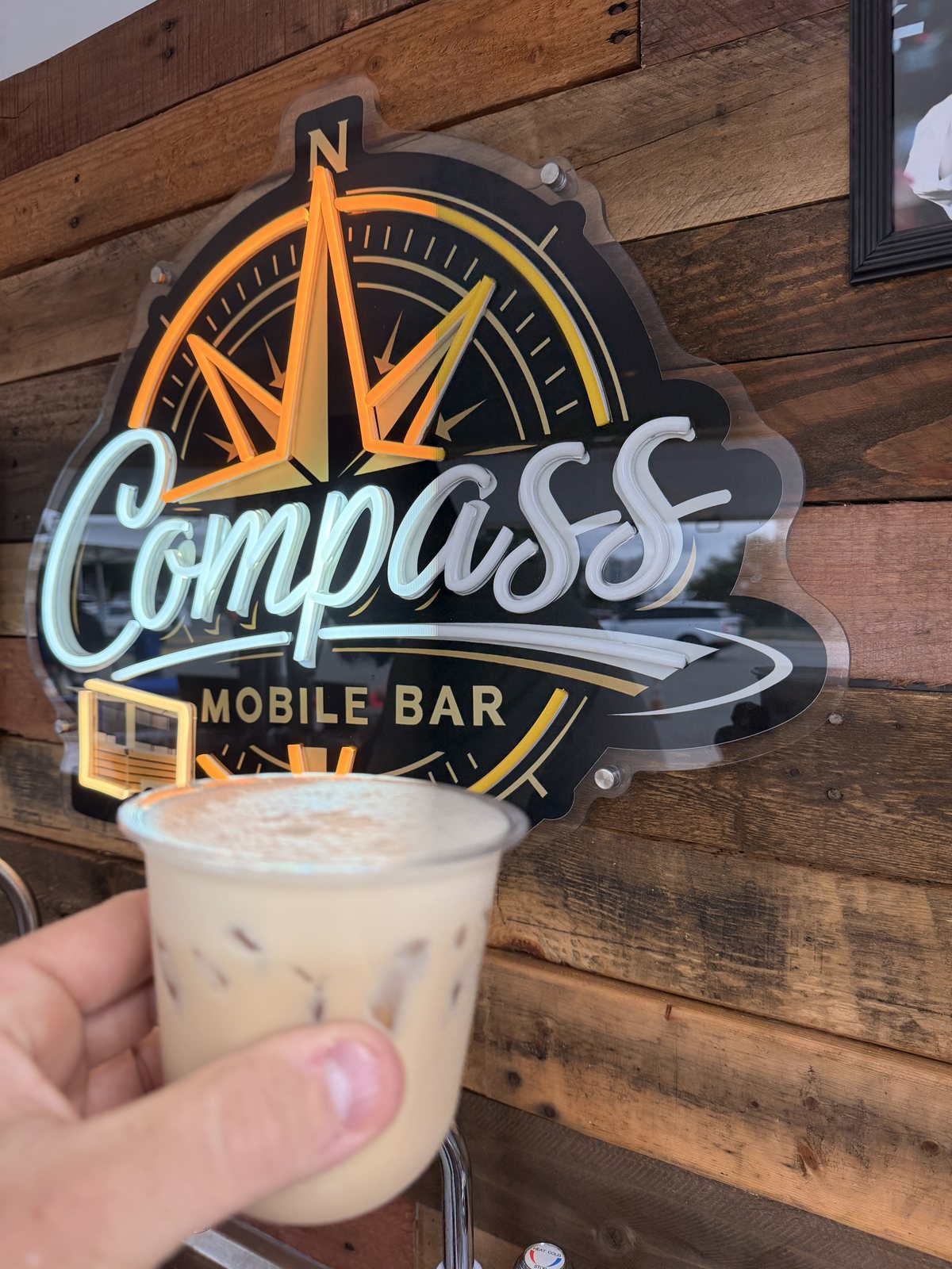 A Kickoff Latte cold brew held up in front of the Compass neon sign